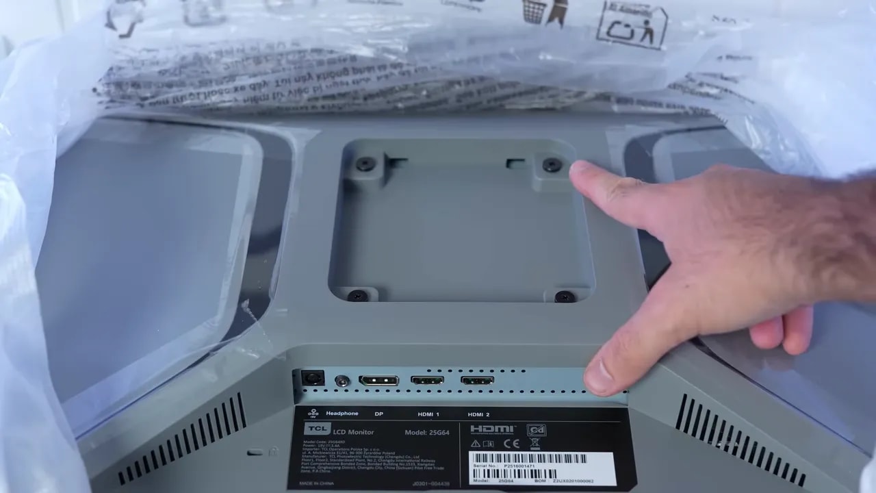 Vista trasera del monitor TCL 25G64 con tapa de conexiones y mano señalando el área de instalación